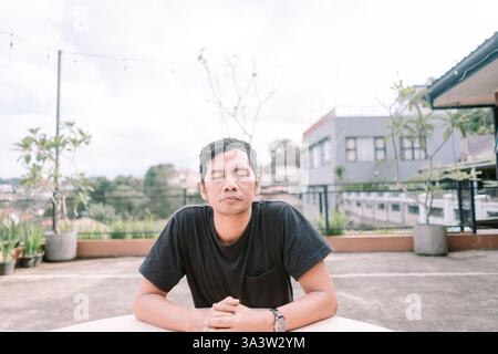 Un uomo asiatico sereno siede a occhi chiusi, praticando consapevolezza e meditazione all'aperto. Egli cerca la pace interiore e uno stato calmo ed equilibrato in mezzo alla u Foto Stock