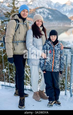 Famiglia di tre persone in piedi in un paesaggio innevato di montagna al tramonto, vestita con caldi abiti invernali, godendo della natura e delle viste panoramiche mozzafiato. Foto Stock