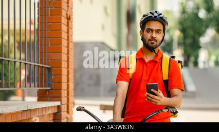 Il cordiale corriere in casco, con zaino, si siede in bicicletta e tiene lo smartphone a portata di mano Foto Stock