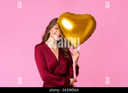 Una donna con un abito marrone regge un palloncino a cuore dorato su uno sfondo rosa Foto Stock