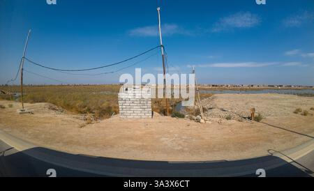 Un paesaggio arido con una piccola struttura in cemento, linee elettriche e una terra asciutta e incrinata. Il cielo è limpido e blu, a indicare una giornata di sole. Nella b Foto Stock