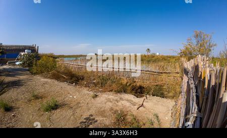 Un tranquillo paesaggio rurale caratterizzato da una recinzione di bambù, un piccolo stagno e vegetazione secca sotto un cielo azzurro. Foto Stock