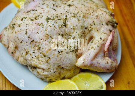 Pollo intero crudo ricoperto di erbe aromatiche provenzali e condimenti, posto su un piatto bianco con fette di limone fresco su un tavolo rustico di legno. Il pollo è preparato per la tostatura, enfatizzando la cucina casereccia e la preparazione dei pasti gourmet. Foto Stock