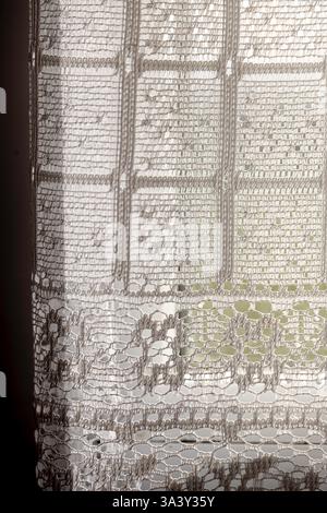 Una tenda in pizzo bianco con un motivo di quadrati e cerchi. La tenda è appesa davanti a una finestra Foto Stock