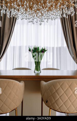 Un'elegante sala da pranzo con un tavolo in legno, sedie trapuntate beige e un vaso di tulipani freschi. Un lussuoso lampadario di cristallo è appeso sopra, completato Foto Stock