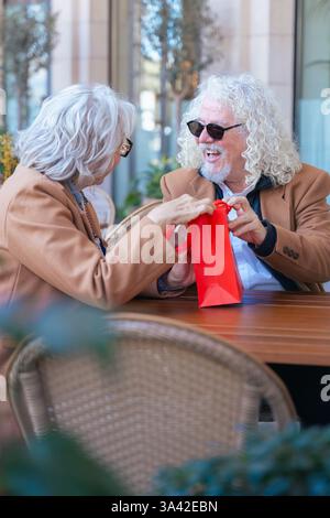 Felice coppia senior caucasica che si scambia un regalo in un caffè all'aperto momento gioioso. Foto Stock