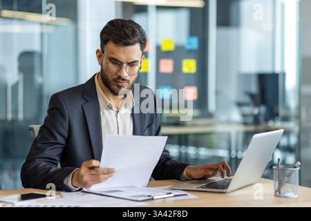 Uomo d'affari serio dietro i documenti, uomo che controlla i rapporti finanziari e di investimento. Finanziere in abiti da lavoro che tiene in mano documenti e documenti, si concentra all'interno dell'ufficio. Foto Stock