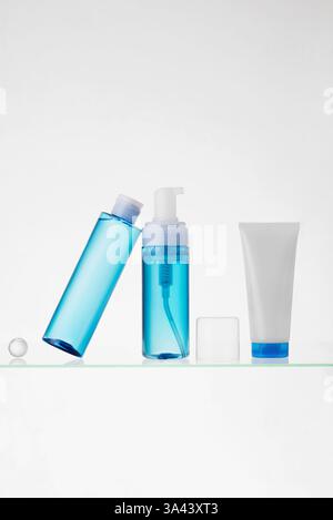 Flaconi blu trasparenti e tubo bianco con prodotti cosmetici su un ripiano in vetro su sfondo bianco. Mockup di prodotti per la cura della pelle Foto Stock