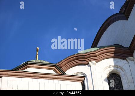 Una chiesa con una croce dorata in cima alla sua cupola, adagiata su un cielo blu vibrante con la luna. Foto Stock