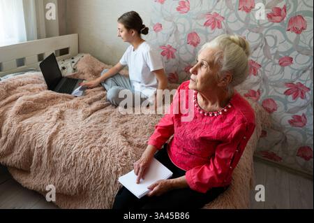 Una giovane donna siede su un letto e lavora su un computer portatile, mentre una donna più anziana osserva in silenzio, catturando un legame sentimentale in una camera arredata in modo confortevole. Foto Stock