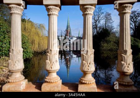 Oldenburg, Germania. 13 marzo 2025. Con il sole, la chiesa dei Lamberti si riflette dietro una storica balaustra nel laghetto con giardino del castello. La balaustra dal design elaborato è una caratteristica del parco storico. Crediti: Hauke-Christian Dittrich/dpa/Alamy Live News Foto Stock
