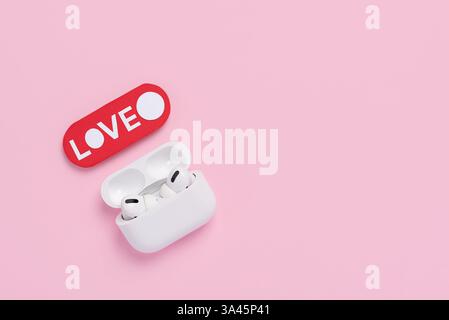 Vista dall'alto degli auricolari bianchi e della parola amore su sfondo rosa pastello. Modello per San Valentino. Concetto di amore e tecnologia, vista dall'alto Foto Stock
