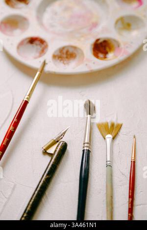 Vari pennelli e strumenti sono disposti su un tavolo di lavorazione accanto a una tavolozza colorata pronta per l'uso. Foto Stock