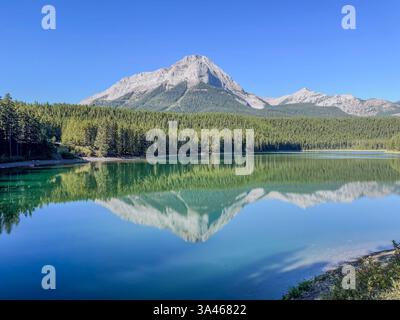 Un tranquillo lago turchese riflette aspre cime di montagna e una fitta foresta sempreverde sotto un cielo azzurro. L'acqua ferma crea uno specchio quasi perfetto Foto Stock