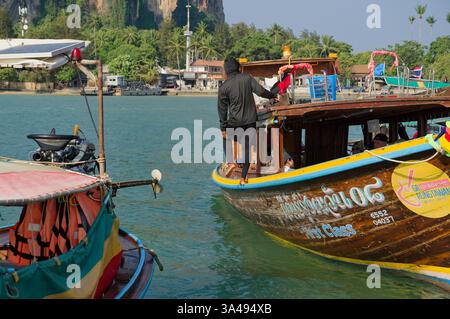 Un operaio di barche mascherato si cala a piedi nudi su una barca di legno a coda lunga al molo galleggiante di East Railay Foto Stock