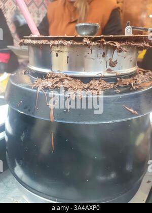 Recipiente rotondo con coperchio per cioccolata calda fusa sul primo piano della parete. Una pentola di cioccolato fuso e molto vapore all'aperto. Pasta dolce al cioccolato. Cibo di strada al festival. Festeggiamenti Foto Stock
