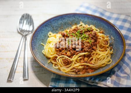 Spaghetti con salsa di carne tritata alla bolognese in un piatto blu, tovagliolo e posate su un tavolo in legno dipinto di bianco, pasto in stile italiano, focalizzazione selezionata, na Foto Stock