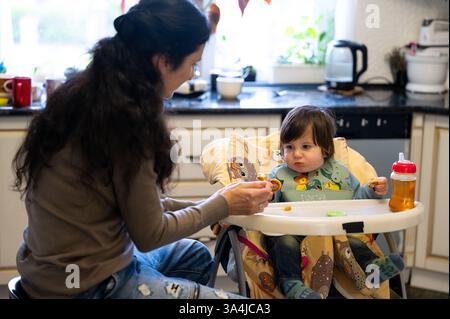 Una madre nutre il suo bambino, che è su un seggiolone, durante il giorno in cucina. Il bambino ha un'espressione carina mentre mangia, rendendolo una preziosa mamma Foto Stock