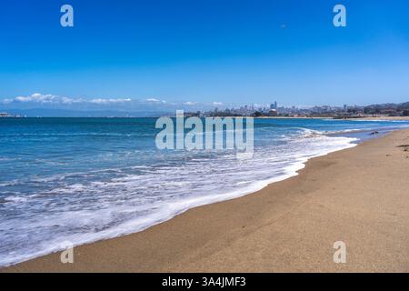 La spiaggia sabbiosa di Golden Gate Beach incontra l'Oceano Pacifico, con lo skyline di San Francisco visibile in lontananza sotto un cielo azzurro Foto Stock
