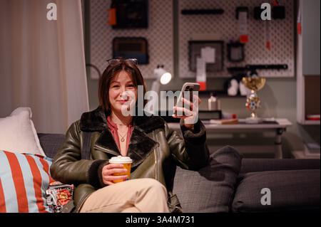 Una donna allegra in una giacca elegante si siede comodamente sul divano, tenendo in mano una tazza di caffè e uno smartphone. Ambiente interno caldo con un'atmosfera accogliente e molto accogliente Foto Stock
