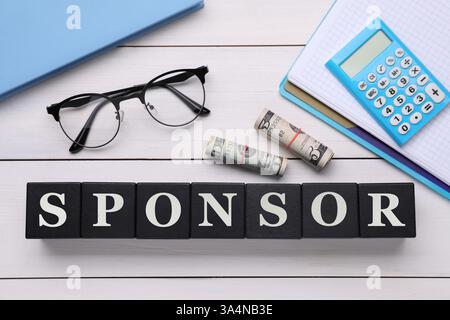 Word Sponsor realizzato in cubi neri con lettere, banconote da un dollaro, occhiali da vista, quaderni e calcolatrice su tavolo in legno bianco, vista dall'alto Foto Stock