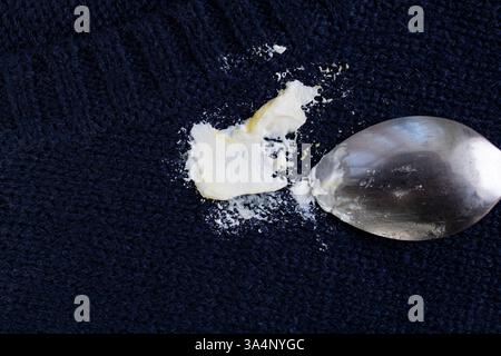 Tessuto in tessuto pullover con una macchia di yogurt bianco e cucchiaio da tè argentato, sfondo astratto Foto Stock