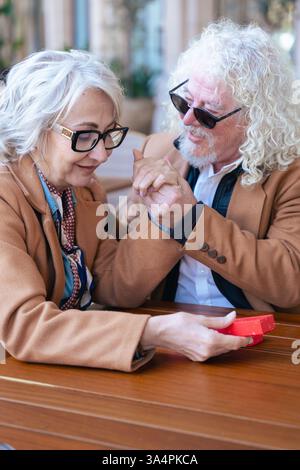 Felice coppia senior caucasica che si scambia un regalo in un caffè all'aperto momento gioioso. Foto Stock
