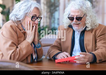 Felice coppia senior caucasica che si scambia un regalo in un caffè all'aperto momento gioioso. Foto Stock