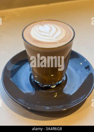 piccolo latte appena fatto con latte art servito in un piccolo bicchiere e piattino, mostrando gli strati della bevanda. Foto Stock