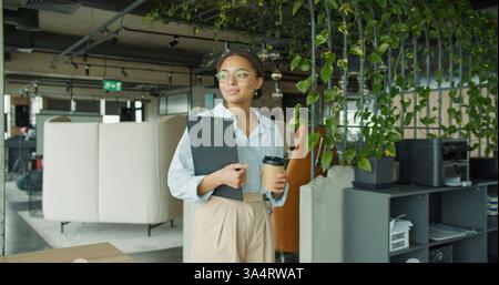 Giovane donna sicura di sé in un ufficio elegante che tiene in mano un portadocumenti e una tazza di caffè, cammina con un sorriso in un moderno spazio di lavoro con piante verdi e. Foto Stock