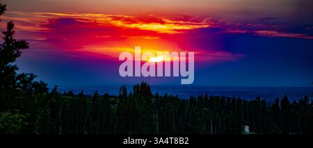 La fotografia cattura un pittoresco tramonto su un paesaggio collinare coperto da fitti alberi. Il sole sta tramontando dietro l'orizzonte, dipingendo il cielo Foto Stock