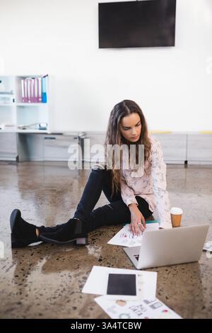 Giovane donna seduta a terra con un laptop e che rivede schizzi di design in ufficio, copia spazio Foto Stock