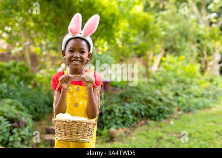 Bambino sorridente che indossa orecchie da coniglietto con cesto con uova in giardino, spazio fotocopie Foto Stock