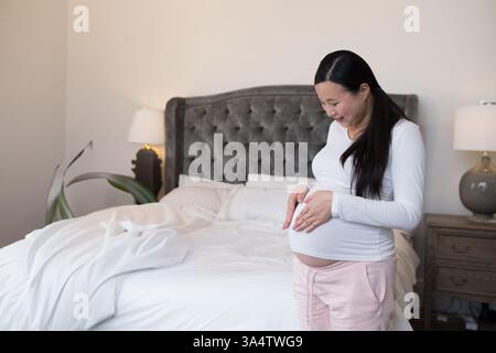 Donna incinta in camera da letto che tocca delicatamente la pancia, sorride di gioia, copia spazio Foto Stock
