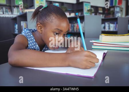 A scuola, una ragazza concentrata che scriveva su un taccuino al tavolo della biblioteca, concentrandosi duramente Foto Stock