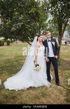 Gli sposi condividono un momento intimo all'aperto al tramonto. La sposa in un velo e un abito bianco che scorre tiene un bouquet di rose bianche come lo sposo in un Foto Stock