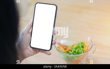 Mani di donna tenere smartphone mock-up schermo bianco sopra dieta perdita di peso cibo fresco e sano. Vista posteriore, copia spazio. Foto Stock