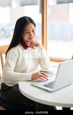 Una donna asiatica sorride mentre guarda lo schermo del notebook, con una mano sotto il mento e l'altra matita in mano. Studiare o lavorare nel bar. Foto Stock