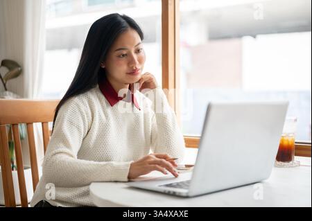 Una donna asiatica guarda con attenzione lo schermo del notebook, con una mano sotto il mento e l'altra con una matita. Studiare o lavorare nel bar. Foto Stock