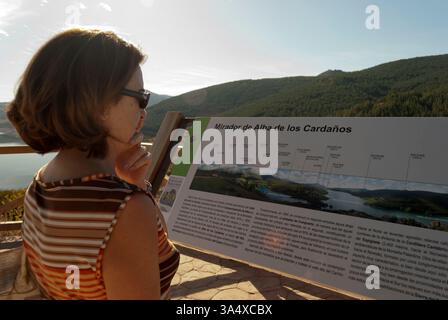 Una donna sta osservando uno spettacolo informativo mentre si gode un punto panoramico naturale che si affaccia sulle lussureggianti montagne verdi e sul tranquillo lago di Alba Foto Stock