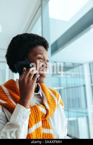 Una donna africana professionista sorridente, con un elegante top arancione, è impegnata in una telefonata all'interno di un ufficio contemporaneo. Incarna fiducia e.. Foto Stock