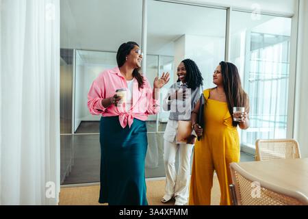 Tre donne professionali si godono una pausa caffè durante una riunione di successo, condividendo sorrisi e chiacchierando. L'immagine cattura un ambiente rilassato ma produttivo Foto Stock