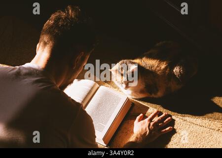 Ragazzo che legge un libro accanto a un gatto. Home ambiente, amore e amicizia tra un umano e un animale domestico Foto Stock