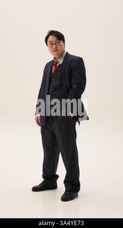 Foto a tutto corpo di un giovane asiatico vestito classico vestito nero e cravatta rossa che guarda la macchina fotografica e posa tenendo le mani in tasca contro il bianco Foto Stock