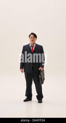Foto a tutto corpo di un giovane asiatico, vestito formale con valigia, si erge pronto per la nuova settimana lavorativa, su sfondo bianco. Foto Stock