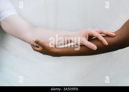 Una donna afroamericana e il suo fidanzato caucasico stanno vicini, toccando le mani vicino a un muro bianco. Questa coppia eterogenea trasmette affetto e connessione adatto a contesti di stile di vita e relazioni. Nessun volto è visibile, il che lo rende versatile per campagne inclusive. Foto Stock