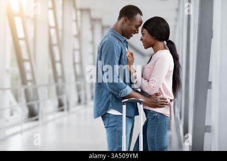 Una romantica coppia afroamericana si abbraccia in un terminal dell'aeroporto mentre si preparano per la loro luna di miele. Un momento di viaggio gioioso, ideale per matrimoni, viaggi o contesti di stile di vita. Perfetto per storie d'amore, viaggi e nuovi inizi. Foto Stock