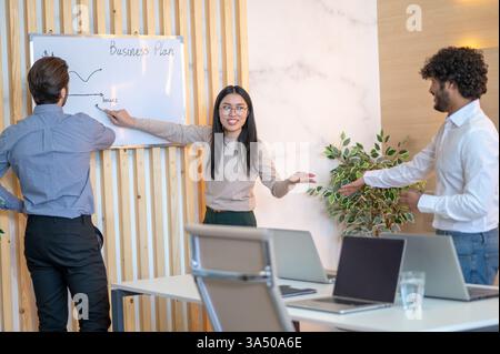 Un dipendente dell'azienda indica un grafico a linee su una lavagna durante una riunione con i colleghi. Questa scena professionale mostra il lavoro di squadra, la pianificazione e la condivisione delle idee in uno spazio di lavoro moderno. Foto Stock