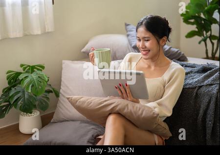 Una donna asiatica attraente si gode il caffè mattutino mentre guarda video su un tablet nella sua camera da letto. La rilassata scena domestica si adatta allo stile di vita domestico, all'intrattenimento digitale e ai temi dell'e-commerce. Ideale per contenuti sul lavoro in remoto, sui contenuti multimediali online o sull'uso quotidiano della tecnologia. Foto Stock