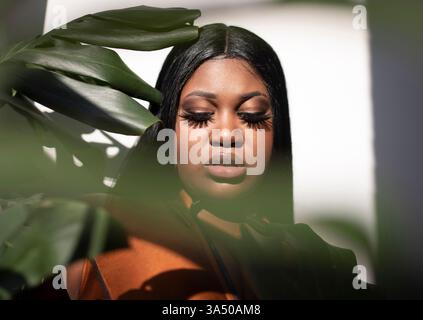 Un modello afro-americano di dimensioni più grandi si trova all'interno, guardando avanti con un'espressione premurosa. Il ritratto enfatizza la sicurezza, la positività del corpo e un ambiente interno moderno. Foto Stock